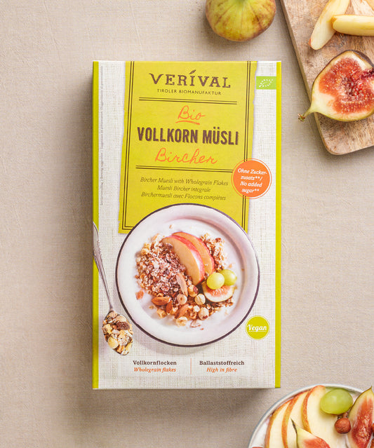Das vegane Bircher Vollkorn Müslis von Verival Bio wird mit Apfelscheiben, Nüssen, mit verstreuten Feigen und einem Löffel auf beigem Hintergrund gezeigt. Es ist reich an Ballaststoffen für einen gesunden Start in den Tag.