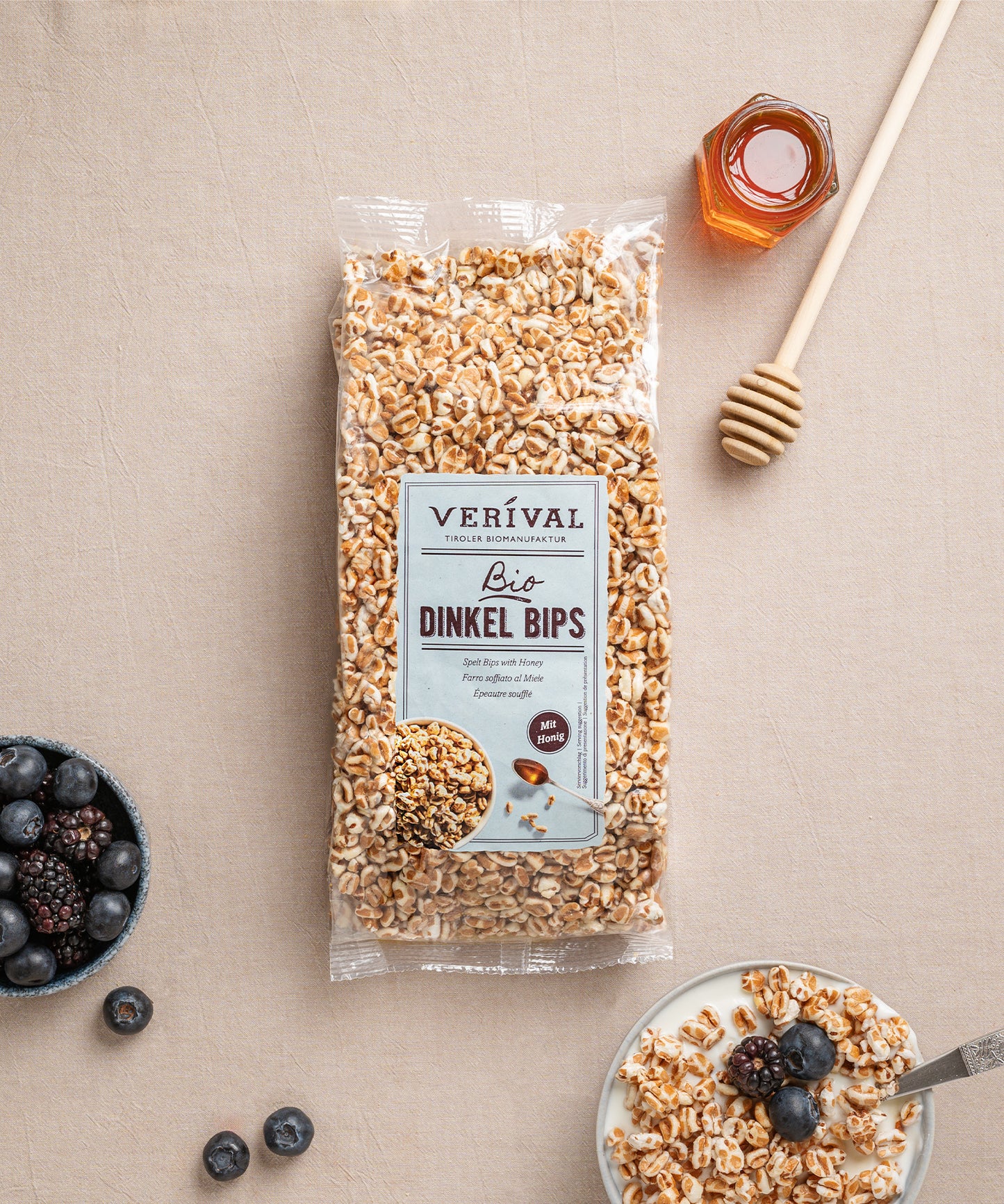 Eine Packung Dinkel Bips mit Honig von Verival Bio steht mit Honig, einem Dipper, prallen Brombeeren, Heidelbeeren und gepufften Dinkelchips in Milch auf einem Tisch.