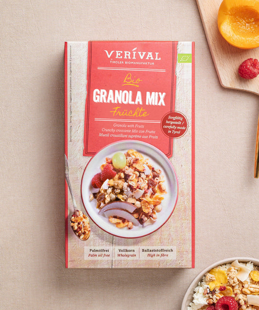 Eine Schachtel Granola Mix Früchte von Verival Bio   steht auf einem beigen Untergrund. Darin befindet sich eine farbenfrohe Schale mit Granola, Nüssen und Trockenfrüchten. Daneben steht eine weiße Schale mit proteinreichem Granola, umgeben von verstreuten frischen Früchten.
