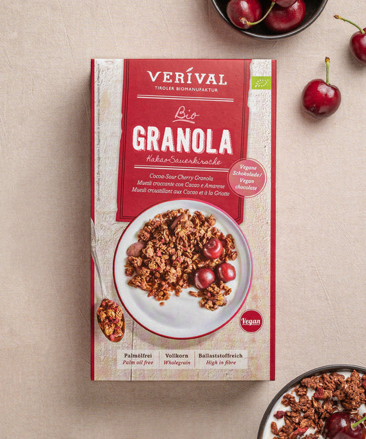 Zu sehen ist eine Produkt aus dem „All I Want For Breakfast“-Set von Verival Bio. Darin befindet sich das Kakao-Sauerkirsch-Granola. Kirschen liegen verstreut herum, und eine kleine Schale mit weiterem Granola ist teilweise sichtbar.