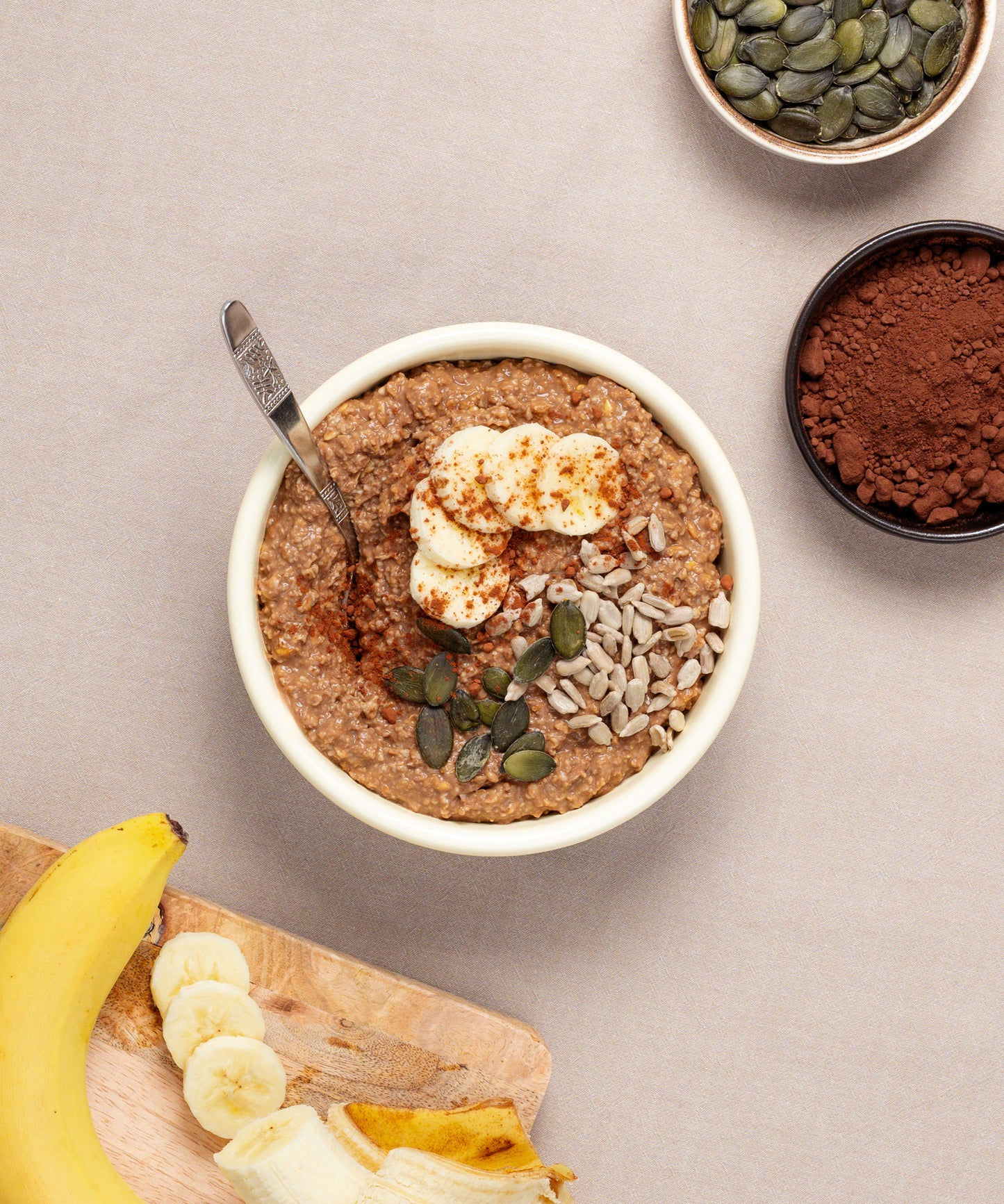 Eine Schüssel Porridge mit Bananenscheiben, Kernen und Kakaopulver lädt zum Verweilen in einer Riess Emaille-Schüssel ein. Ein Löffel liegt darin, während ein kleines Schälchen mit zusätzlichen Kernen und Kakao auf einem Holzbrett bereitsteht, um Ihr Frühstücksritual zu verfeinern.