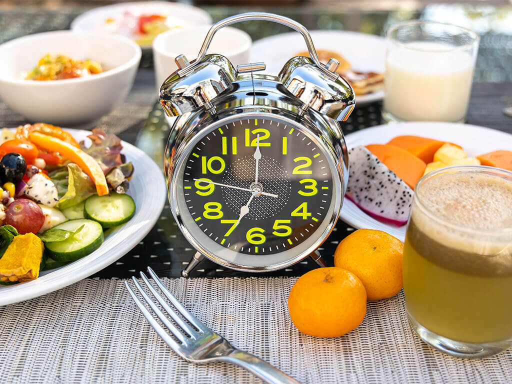 A silver analog alarm clock stands on a table among various breakfast items, including salad, fruit, juice, and a glass of milk.