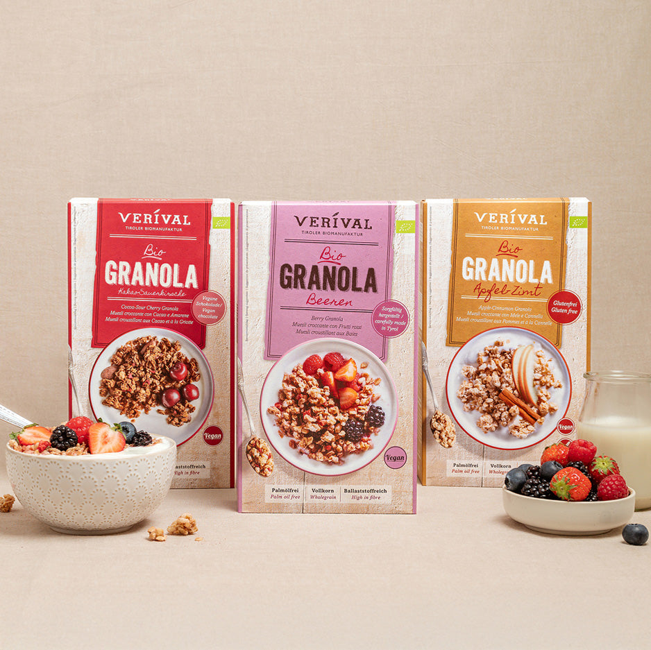 Three boxes of Verival wholegrain muesli in different flavors are presented with a bowl of muesli, a glass of milk and various berries on a beige background.