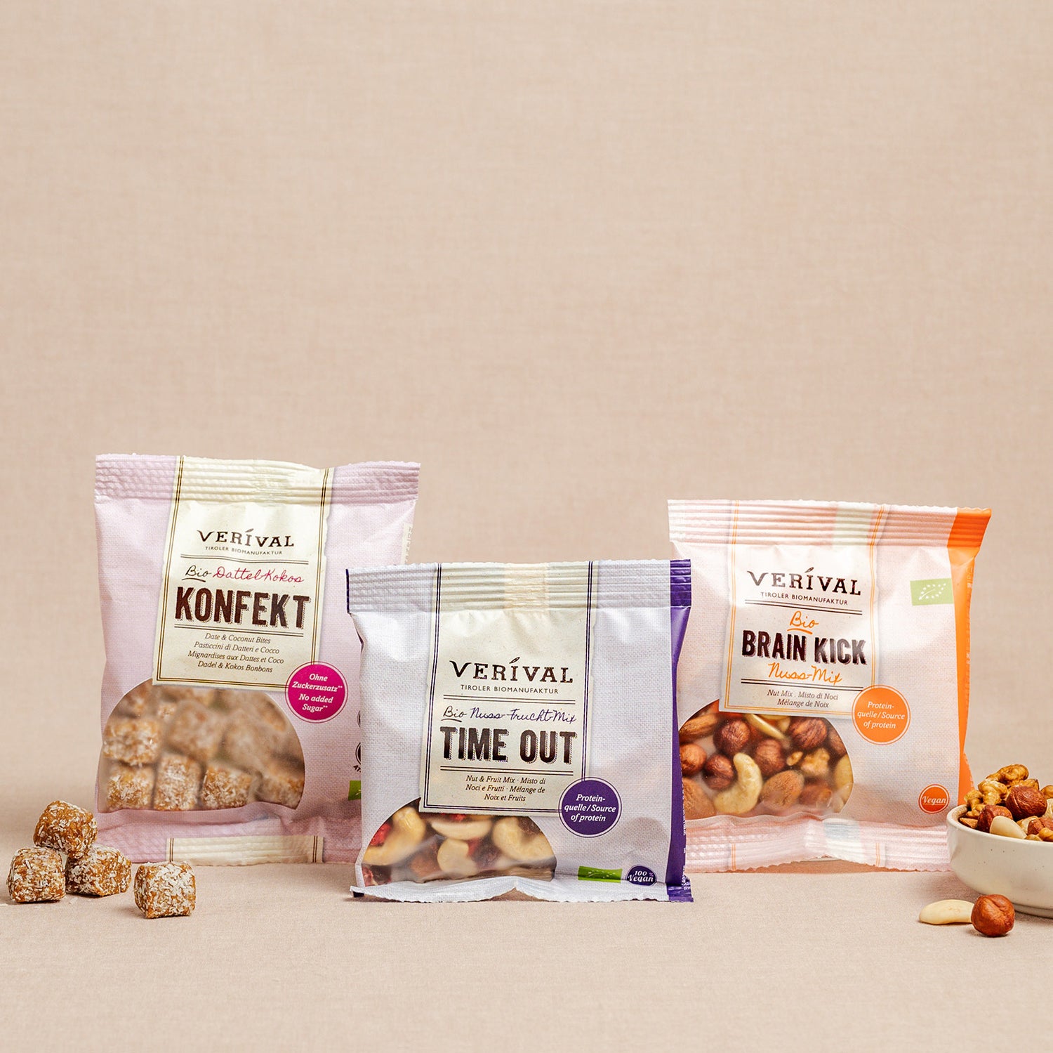 Three Verival snack packs – Konfekt, Time Out and Brain Kick – are presented upright on a neutral surface, with some of their contents scattered nearby.