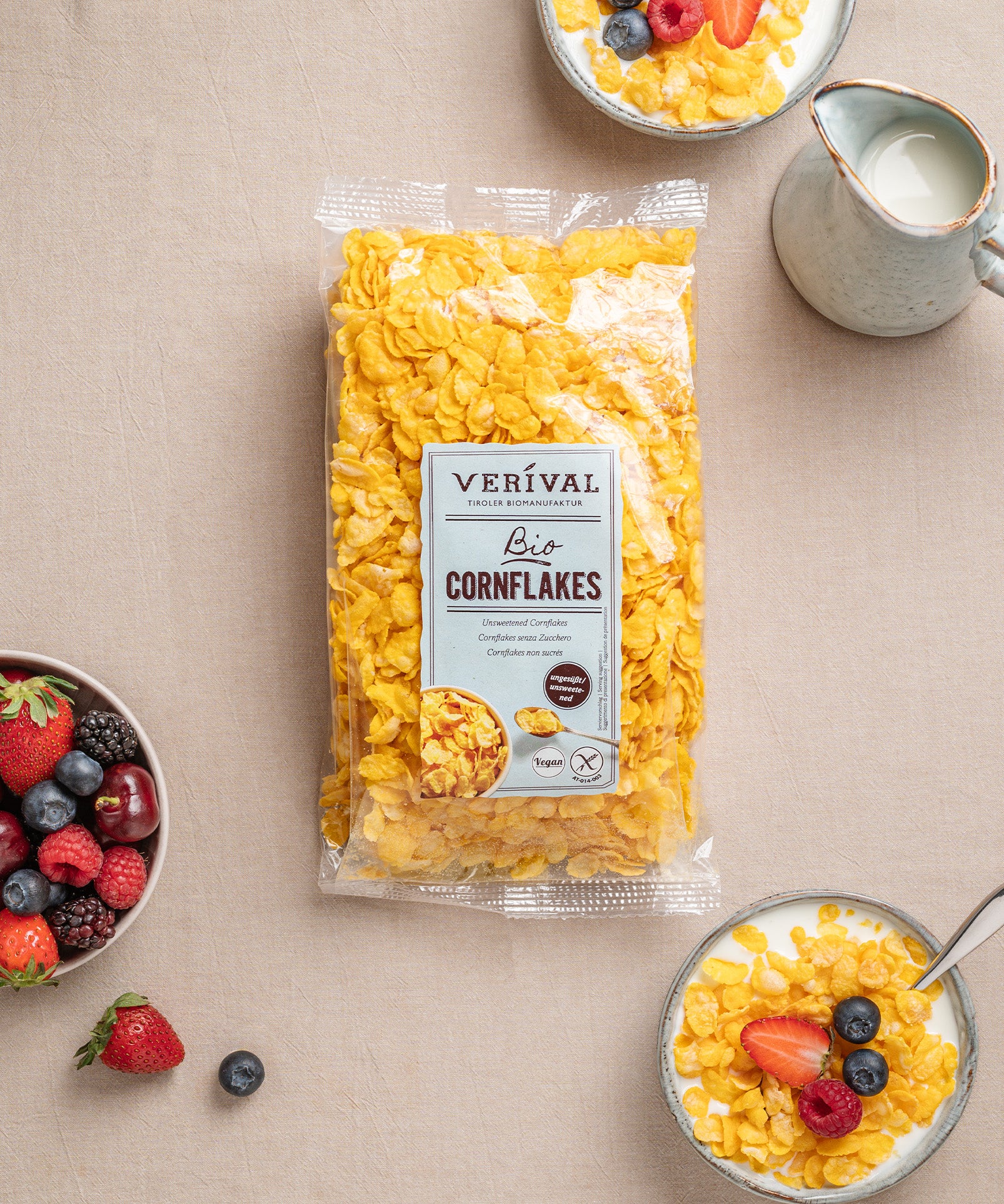 Eine Packung ungesüßte, knusprige und leckere Cornflakes von Verival Bio steht zwischen Müslischalen mit Milch, Beeren und einem Krug, elegant arrangiert auf einer beigen Tischdecke.