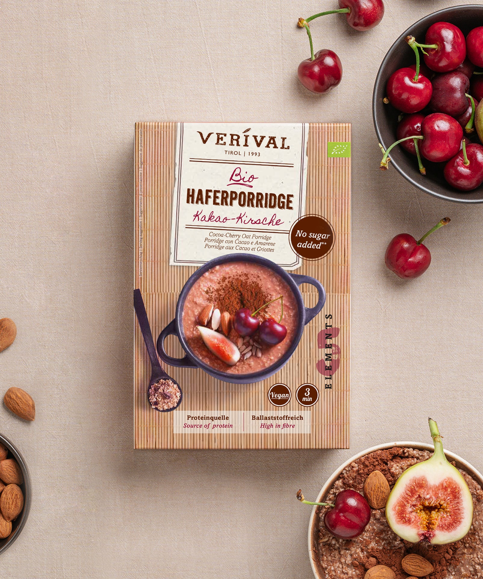 Eine Schachtel Verival Bio GmbH Bio Haferporridge Kirsche-Kakao steht auf einem hellen Tisch, umgeben von Kirschen, Feigen, Mandeln und einer Schüssel mit ballaststoffreichem Porridge - perfekt für ein warmes Frühstück.