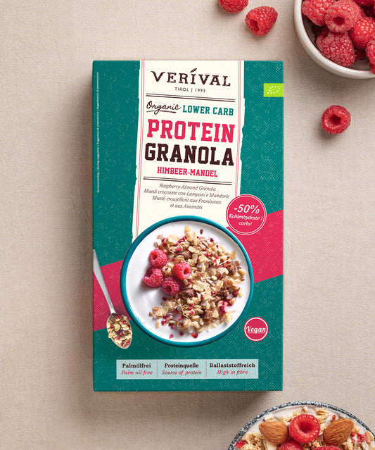 Eine Packung „Bio Protein Lower Carb Granola Himbeer-Mandel“ der Verival Bio GmbH steht auf einer hellen Unterlage. Auf der Verpackung werden die Eigenschaften „vegan“, „pflanzliches Protein“, „ohne Palmöl“, „proteinreich“ und „ballaststoffreich“ hervorgehoben. Daneben stehen Schalen mit Granola, Himbeeren und einem Löffel.