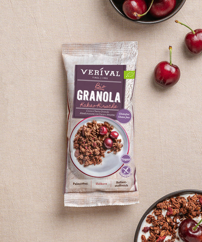 Eine Packung Verival Bio GmbH Bio Kakao-Kirsche Granola glutenfrei, eine vegane und ballaststoffreiche Leckerei, steht auf einem Tisch mit frischen Kirschen und Schalen mit glutenfreiem Knuspermüsli und Kirschen daneben.