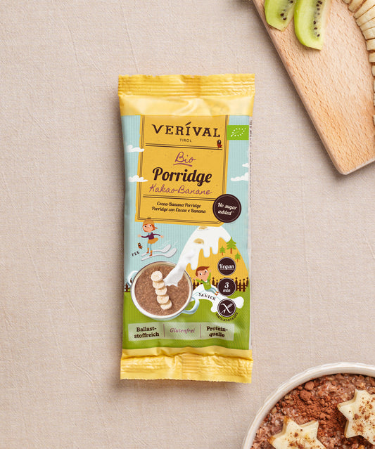 Eine Packung Verival Bio GmbH Bio Kids Porridge Kakao-Banane liegt auf beigem Untergrund neben einem Holzbrett mit Kiwi-Scheiben und einer Schale Porridge mit Bananenscheiben.